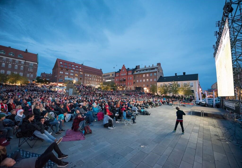 Människor sitter på torget och ser på en storbildsskärm