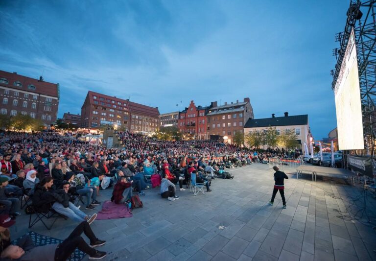 Människor sitter på torget och ser på en storbildsskärm